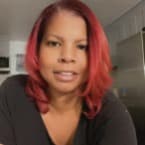 Woman with vibrant red hair, wearing a black top, smiles warmly at the camera. A bright kitchen setting is visible in the background.