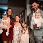 A family of six poses for a photo: a mother in a red dress holding a baby in white, a father in a gray suit holding a baby in white, and two young girls in floral dresses standing between them.