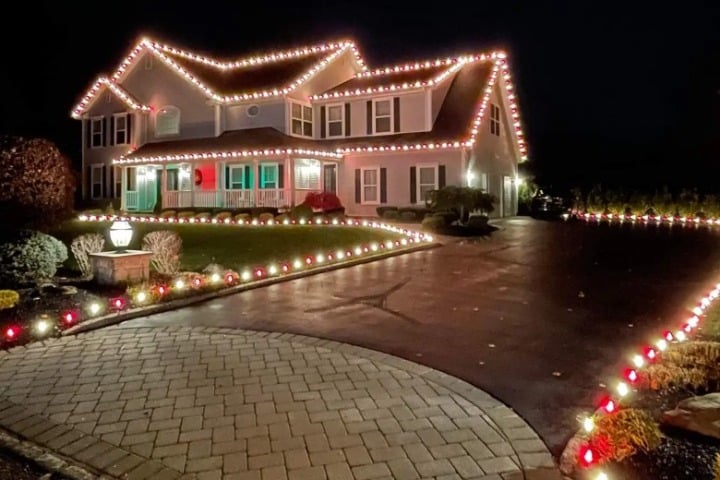 A two-story house is decorated with festive Christmas lighting. Lights line the roof, porch, and driveway, creating a warm, inviting holiday scene.