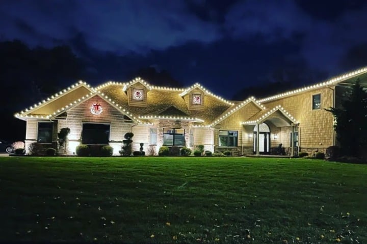 Large house adorned with warm white Christmas lighting along the roofline. Wreaths hang above the windows, set against a dark, cloudy night sky.