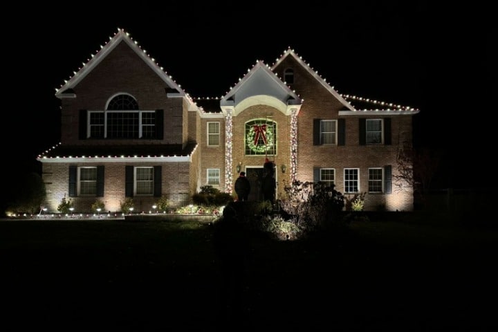 Brick house adorned with Christmas lighting, highlighting rooflines and landscaping. A large wreath hangs above the entryway, creating a festive holiday scene.