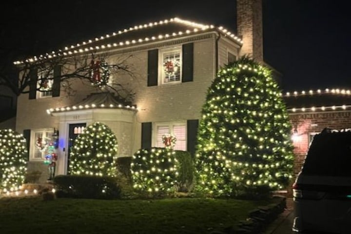 Festive Christmas lighting adorns a two-story house and its landscaping. Warm white lights outline the roof, windows, and wrap the bushes, creating a cozy holiday glow.