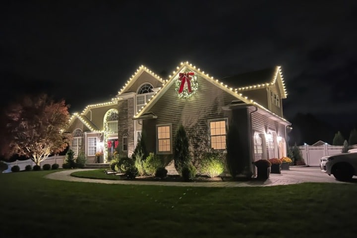 House decorated with Christmas lights, outlining the roof and gable peaks. Wreaths with red bows accent the front. Festive holiday display at night.