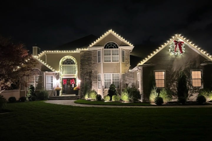 Elegant home featuring Christmas lighting outlining the roofline. Wreaths with red bows adorn the entrance and a gable, adding festive cheer.