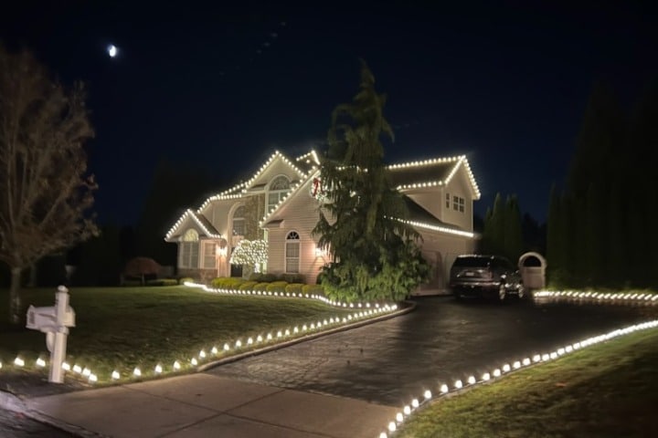 Suburban home aglow with Christmas lighting. White lights line the roof and driveway, creating a festive holiday scene under a dark, moonlit sky.