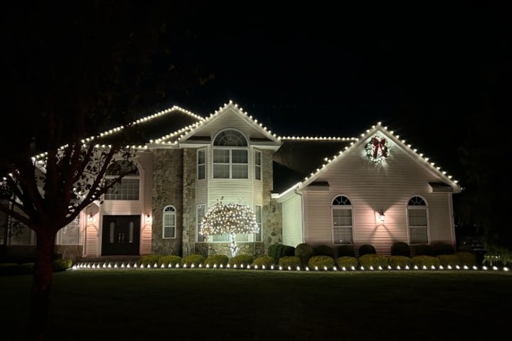 Elegant home adorned with warm white Christmas lighting outlining the roofline, windows, and landscaping. A festive wreath with a red bow hangs above the garage.