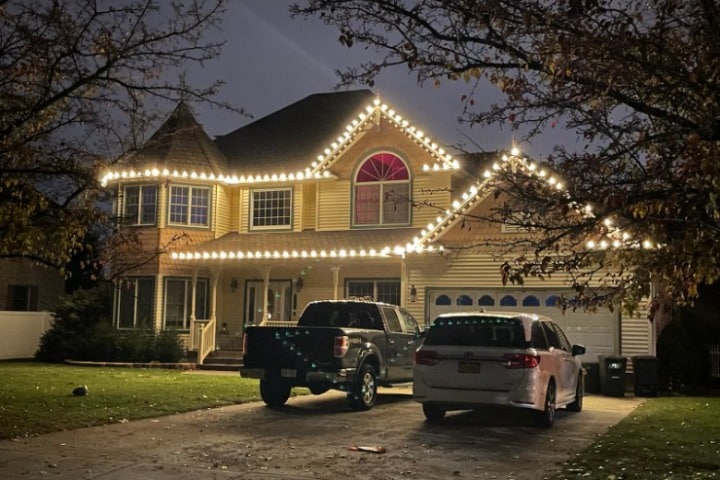 Suburban house adorned with bright Christmas lighting, outlining the roofline and windows. A black pickup truck and white minivan are parked in the driveway, adding to the festive holiday scene.