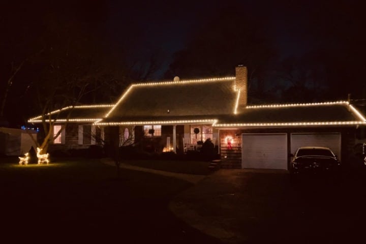 House decorated with warm white Christmas lighting along the roofline. Two illuminated reindeer stand on the lawn, with a dark car parked in the driveway.