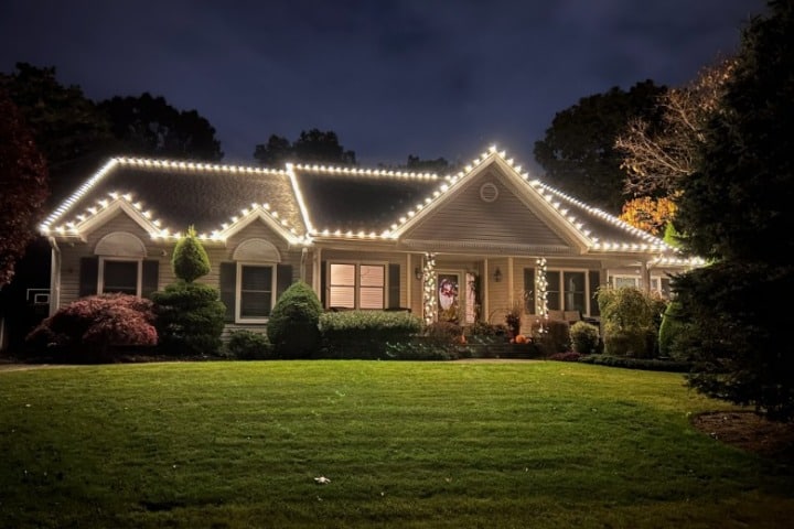 A suburban home glows at night, outlined with warm white Christmas lights along the roofline. Landscaped yard and a wreath on the front door add to the cozy, festive atmosphere.