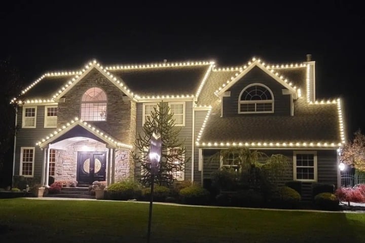 Large two-story house adorned with warm white Christmas lights outlining the roofline. The home features a stone facade, dark gray siding, and manicured landscaping, creating a festive and inviting holiday scene.
