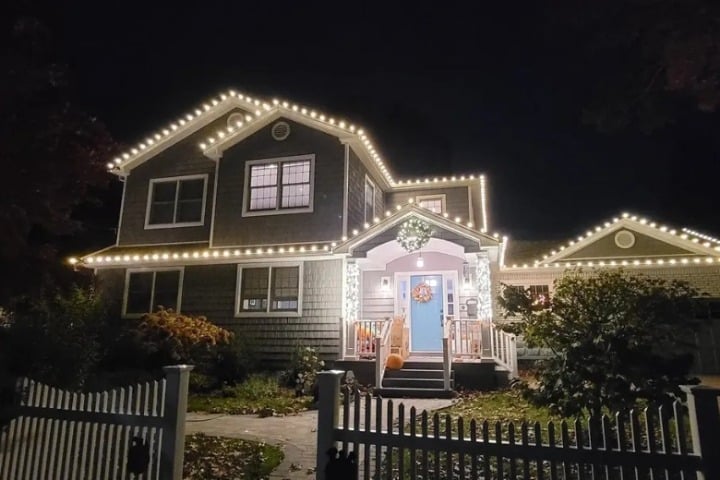A two-story house is decorated with warm white Christmas lighting outlining the roofline. A wreath hangs above the blue front door, with a white picket fence in the foreground.