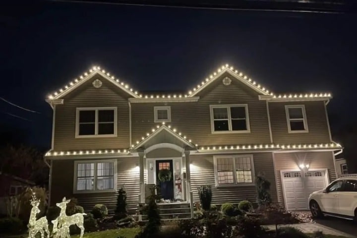 A two-story house is decorated with bright white Christmas lighting outlining the roof. Illuminated reindeer stand on the lawn. A wreath hangs on the front door, creating a festive holiday scene.