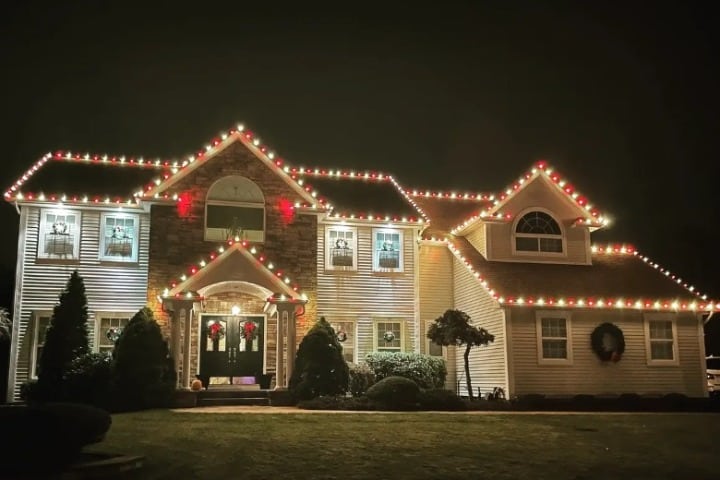 Festive two-story house adorned with red and white Christmas lighting along the roofline. Wreaths hang on the front door and windows, creating a warm, inviting Christmas Lighting display.