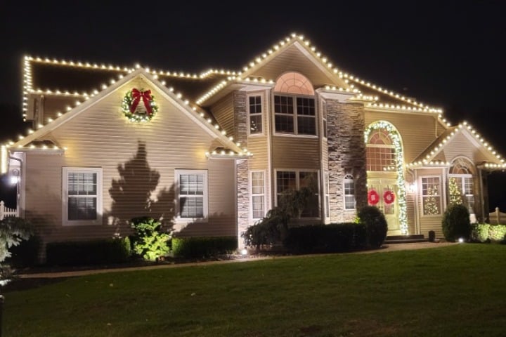 Large house decorated for Christmas with warm white lights outlining the roofline and wreaths on the front doors. The lawn is green and well-manicured.