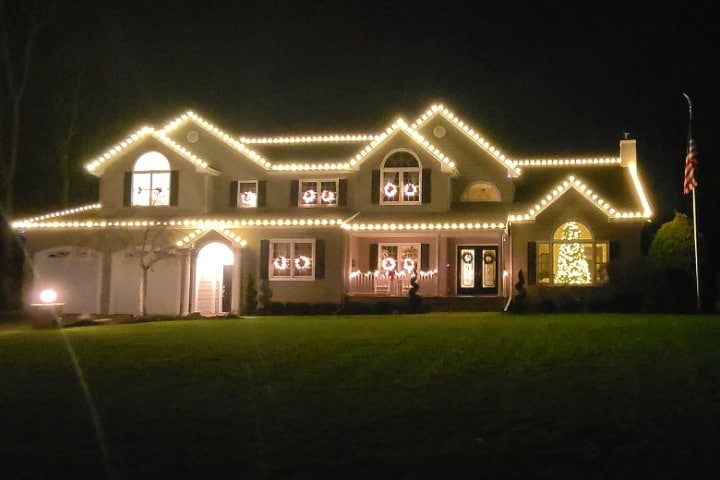 A large home is decorated with Christmas lighting. Warm white lights line the roof, and wreaths adorn the windows. A lighted Christmas tree is visible through a front window.