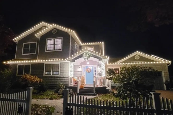 Home featuring Christmas lighting: a charming gray house is outlined with bright white Christmas lights, highlighting the roofline. A blue door with a wreath adds a festive touch. White picket fence in the foreground.