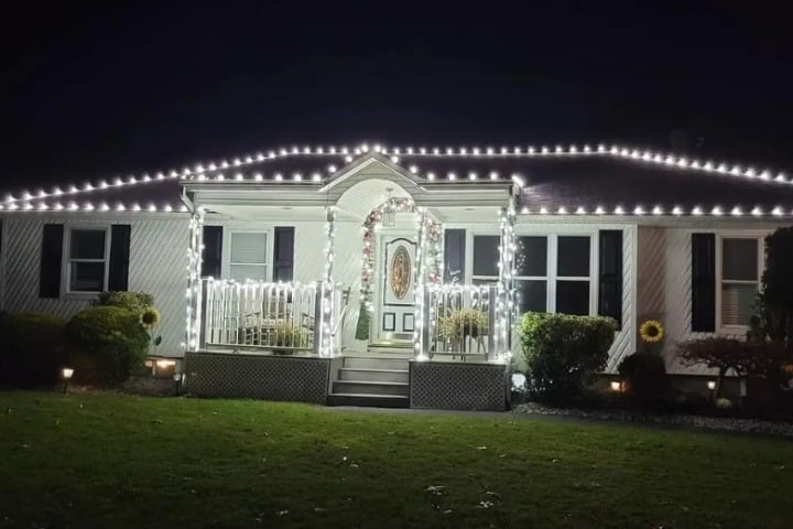 Home adorned with Christmas lighting. White lights outline the roof, porch, and doorway, creating a warm, festive glow. Landscaped lawn and shrubs add to the charming holiday scene.
