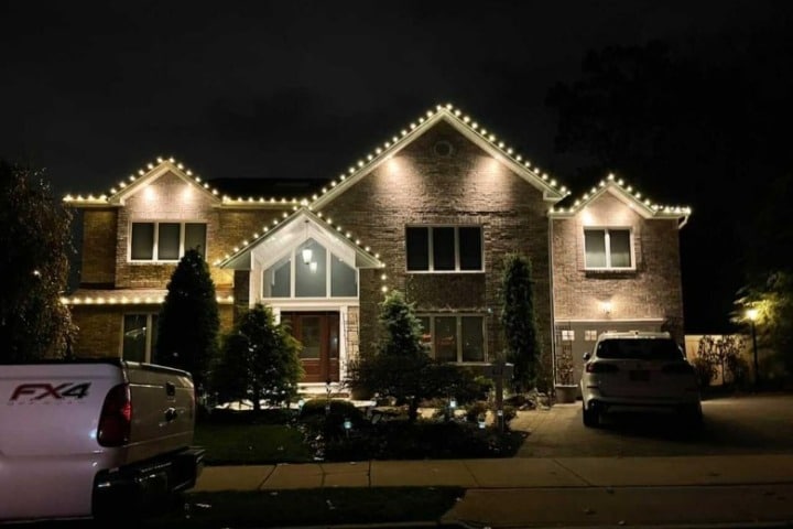 Brick house at night illuminated with warm white Christmas lights outlining the roofline. A white pickup truck is parked in the driveway.