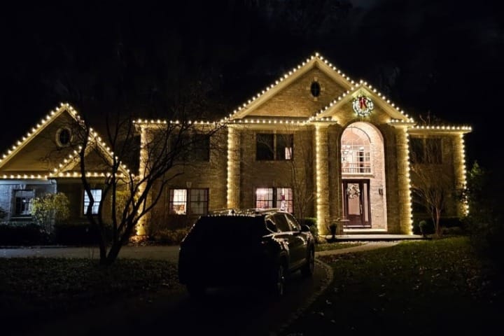 Large stone house decorated with warm white Christmas lights outlining the roofline. A dark SUV sits in the driveway. A wreath hangs above the front door.