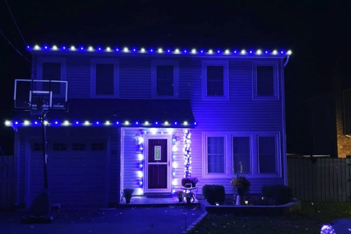 Two-story home decorated with blue and white Christmas lighting outlining the roof and doorway. A basketball hoop stands to the side of the garage.