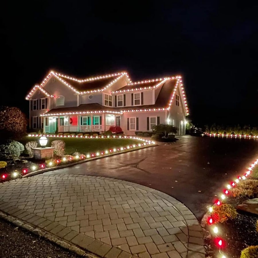 Festive Christmas lighting adorns a large two-story house at night. Red and white lights outline the roof, porch, and driveway, creating a warm, holiday glow.