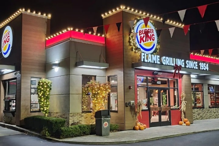 Burger King restaurant decorated with Christmas lighting and fall decor. The sign reads "Burger King, Flame Grilling Since 1954." Pennant banners and string lights adorn the roofline.