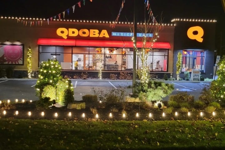 Qdoba restaurant brightly decorated with commercial holiday lighting at night. String lights line the roof and landscape, creating a festive atmosphere.