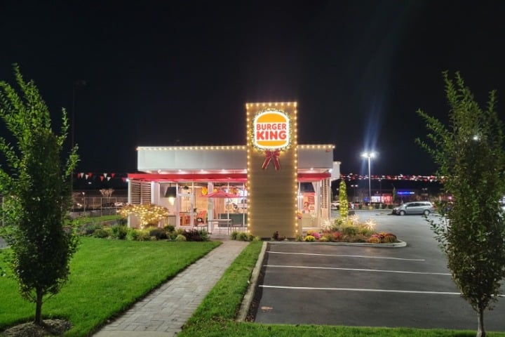 Burger King restaurant illuminated with commercial holiday lighting at night. The building is outlined with warm white lights, enhancing its festive appearance. Well-lit parking lot and landscaping add to the inviting atmosphere.