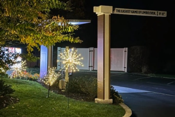 Illuminated holiday decorations sparkle in front of a commercial building at night. A sign reads "The Easiest Game of Limbo Ever! 9'-0". Commercial holiday lighting enhances the building's entrance.