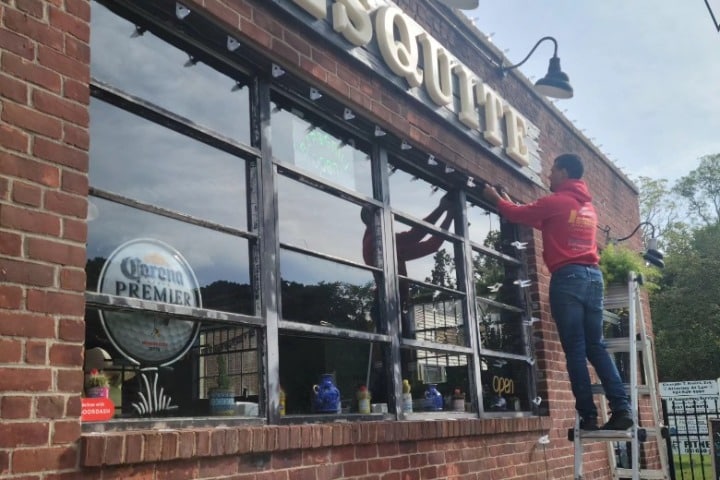 A worker installs commercial holiday lighting on a brick building with large windows. The sign "Mesquite" is visible above the windows.