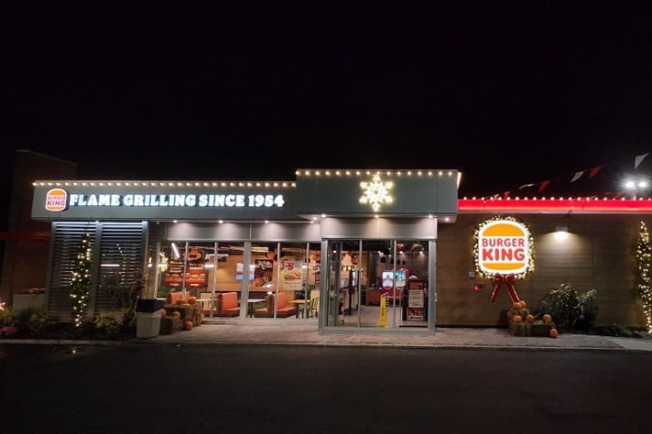 Burger King restaurant at night, decorated with commercial holiday lighting, including string lights and a snowflake. Pumpkins and hay bales add a festive touch to the exterior.