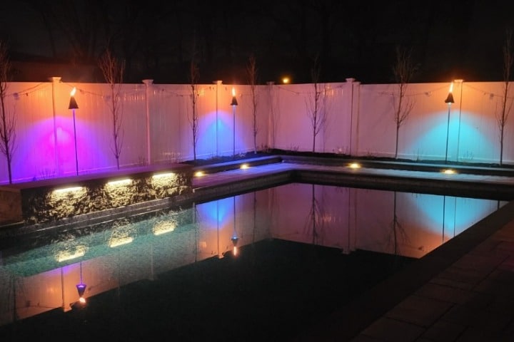 Nighttime landscape lighting illuminates a backyard pool. Tiki torches cast colorful light on a white fence, reflected in the still water. Stone accents and subtle in-ground lights add to the ambiance.