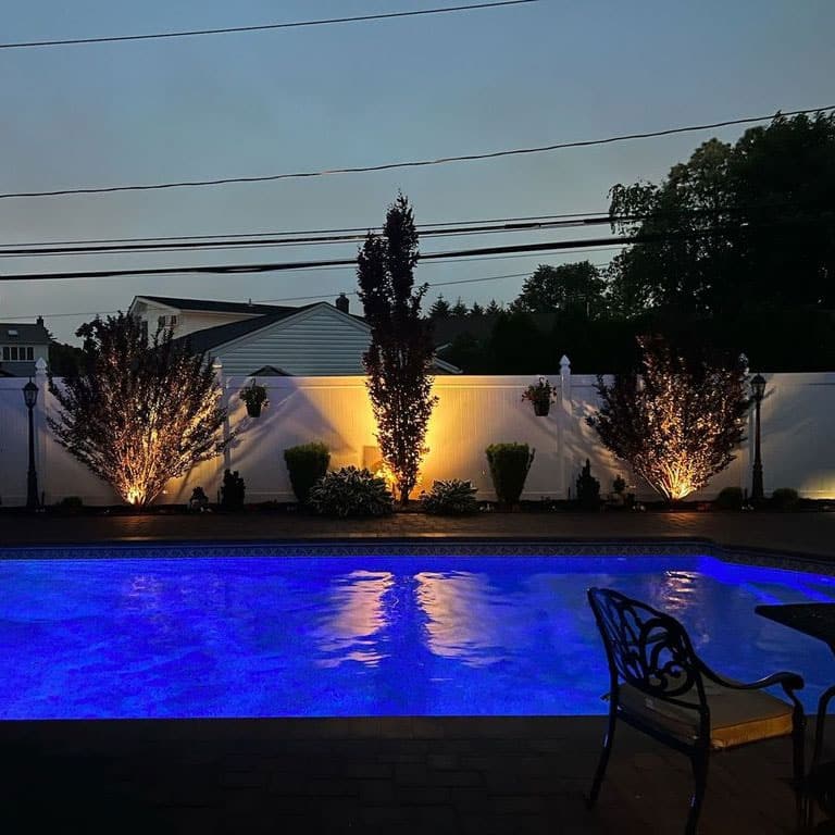 Backyard pool with blue lighting reflecting on the water, surrounded by landscape lighting illuminating trees and bushes against a white fence at dusk.