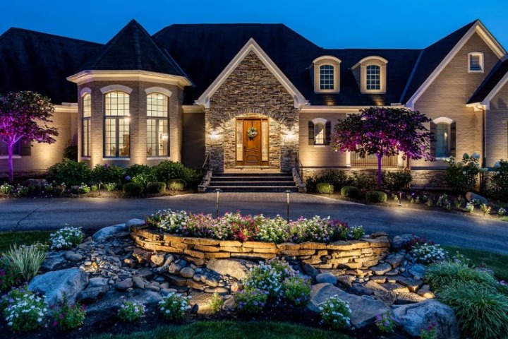 Elegant home illuminated with landscape lighting at dusk. Stone facade, manicured landscaping, and a water feature create a welcoming entrance.