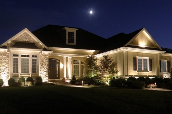 Elegant home illuminated by landscape lighting at night, showcasing architectural details and manicured lawn under a full moon.