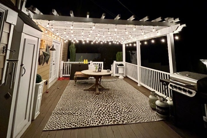 Outdoor deck at night with Christmas lighting strung across a white pergola. A round table and patterned rug sit on the wooden deck next to a grill, creating a cozy home atmosphere.