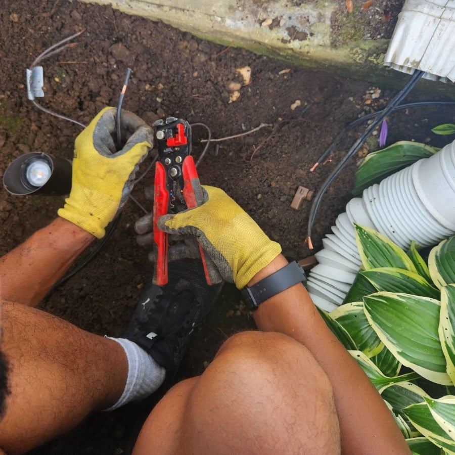 Installing landscape lighting: A person kneels outdoors, connecting wires for landscape lighting with wire strippers near a white downspout and hosta plants.