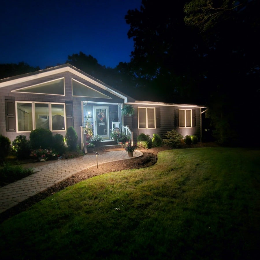 Twilight view of a single-story home illuminated with landscape lighting. The walkway is lit, leading to a porch with a rocking chair, with soft light accentuating the house and yard.