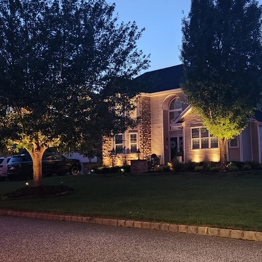 Suburban home at dusk featuring landscape lighting that highlights architectural details and surrounding trees. Lights accent the stone facade and manicured lawn.