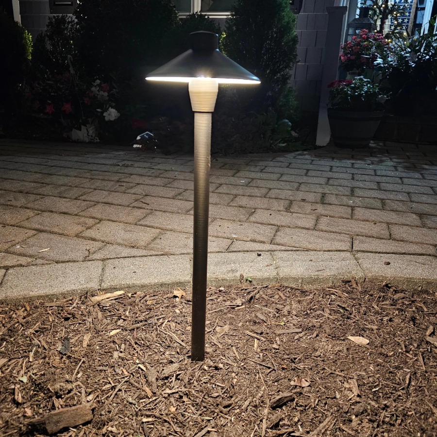 A landscape lighting fixture illuminates a mulch-covered garden bed at night. The light shines down onto the brick walkway, highlighting the surrounding greenery.
