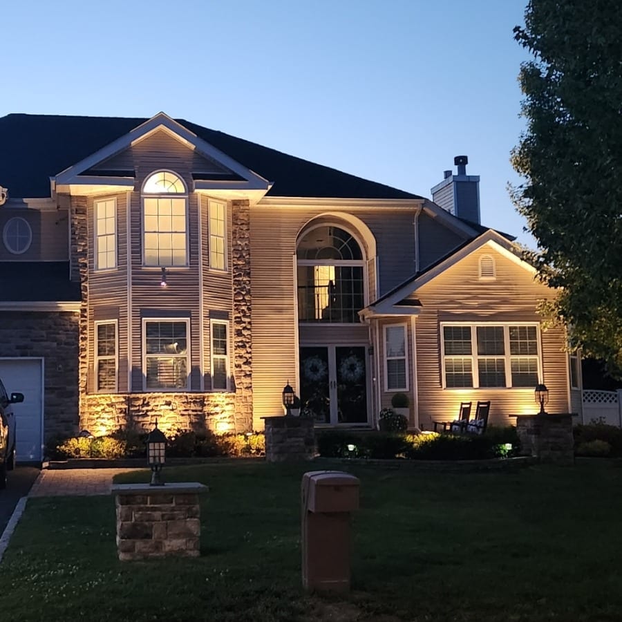 Elegant home illuminated with landscape lighting at dusk. Warm lights highlight architectural details and stonework, creating a welcoming ambiance.