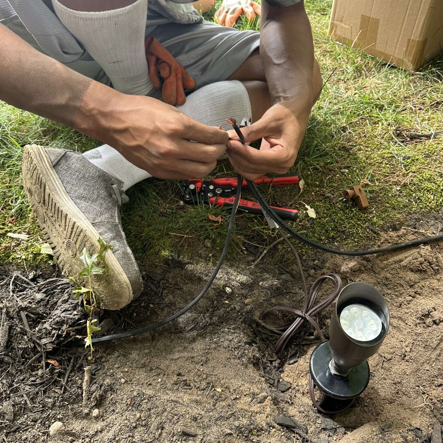 Installing landscape lighting: a person connects wires from a spotlight, with wire strippers and orange gloves nearby on the grass.