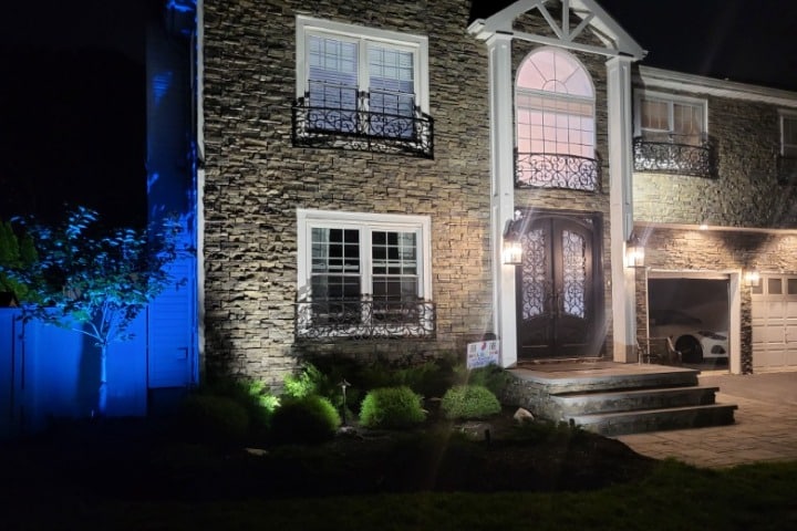 Elegant stone facade of a two-story home at night, illuminated by warm spotlights and cool blue accent lighting on a tree. Ornate wrought iron balconies adorn the windows above the front door.