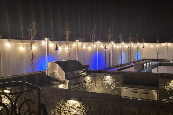 Outdoor kitchen and landscape lighting at night. String lights adorn a white fence with trees behind it, illuminated by blue uplighting. A built-in grill and counter space are lit by warm, recessed lights.