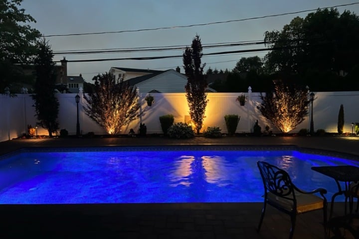Backyard pool at dusk with landscape lighting illuminating the white fence and surrounding trees, creating a serene and inviting atmosphere. The pool water glows blue.