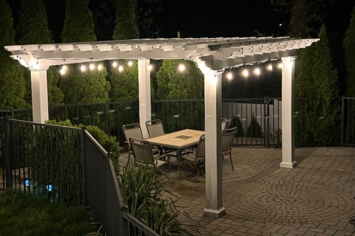 Outdoor patio with a white pergola illuminated by string lights at night. The scene showcases landscape lighting highlighting a dining table and chairs on a brick patio.