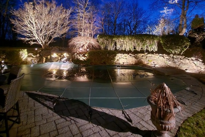 Nighttime view of a covered pool with outdoor lighting illuminating the surrounding trees and stone wall in a cozy backyard. This home's landscape is beautifully enhanced with strategic lighting.