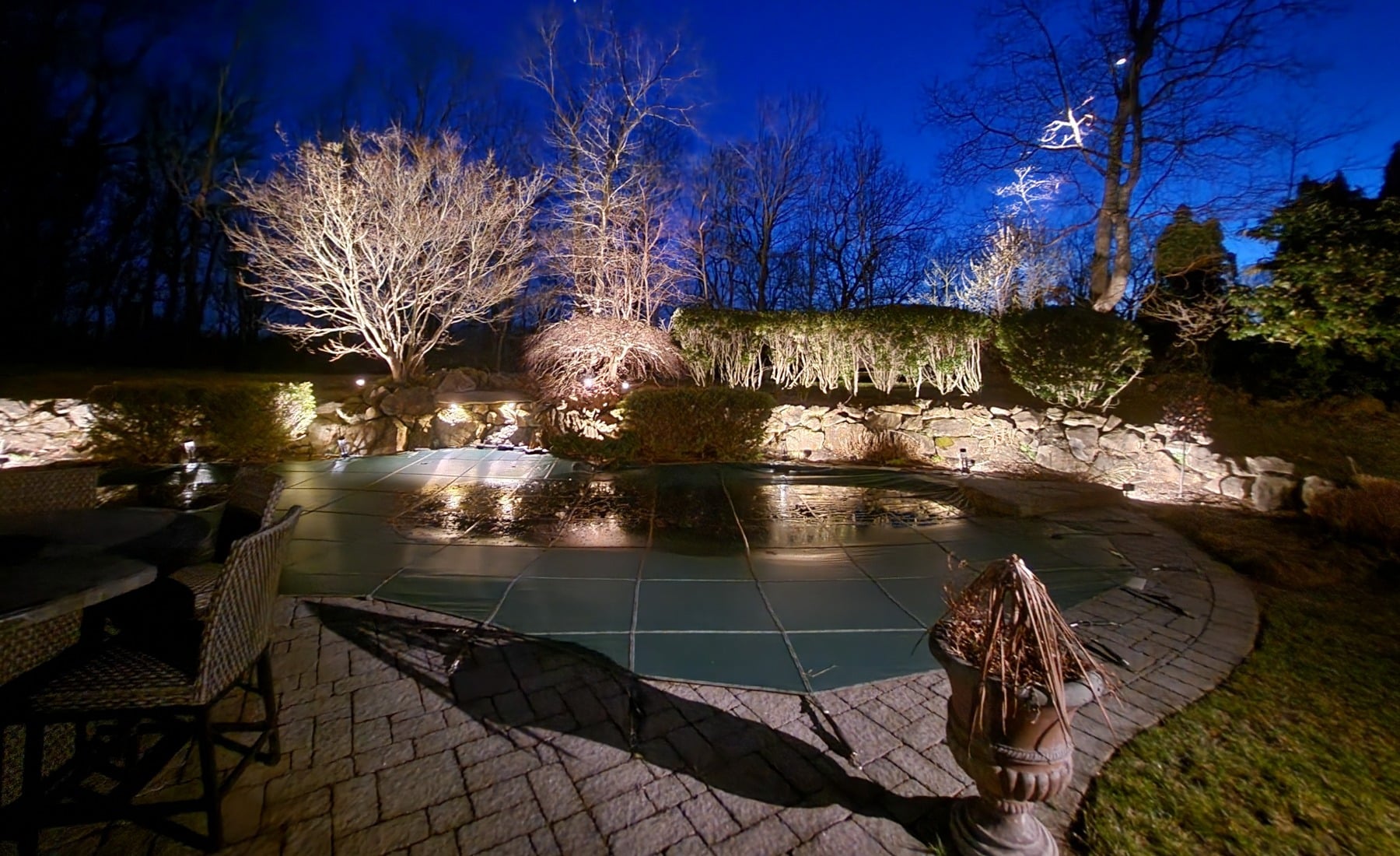 Backyard landscape lighting illuminates a covered pool at night. Warm lights highlight trees and stone walls, creating a serene and inviting outdoor space.