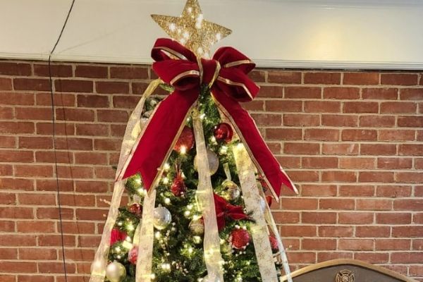 A Christmas tree is decorated with red ornaments, gold ribbon, and a large red bow, topped with a gold star. The Christmas lighting illuminates the tree against a brick wall backdrop.