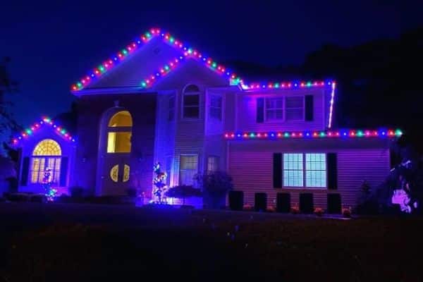 A two-story house is adorned with colorful Christmas lighting outlining the roofline and windows, set against a dark night sky.